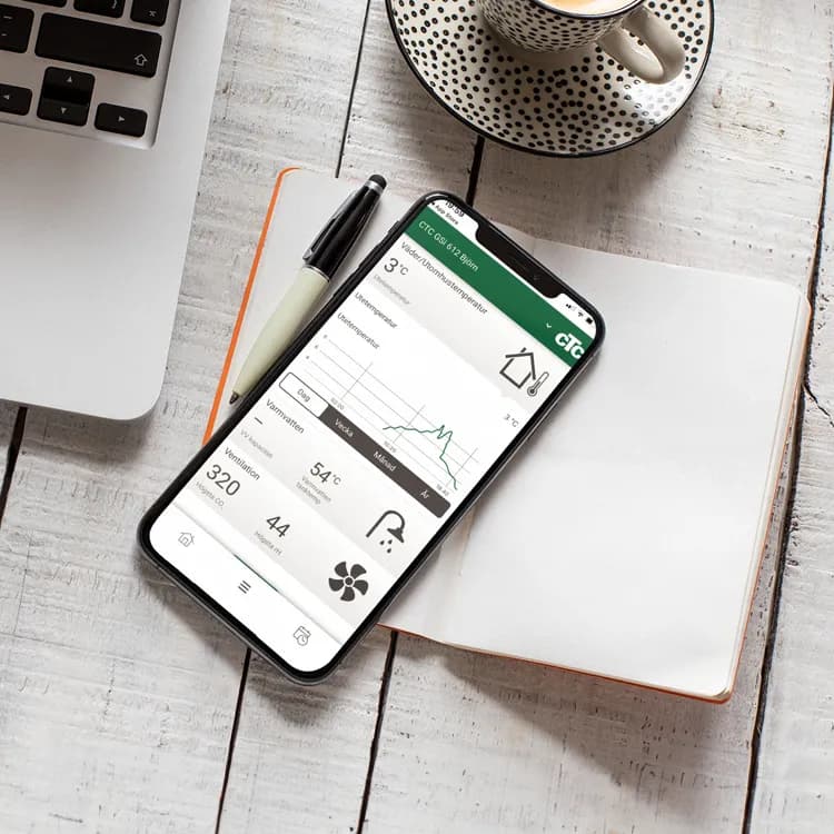 En smartphone på en öppen anteckningsbok visar en väderapp, med en penna vilande i närheten. I bakgrunden står en bärbar dator redo och en kaffekopp ångar försiktigt, vilket skapar förutsättningar för en rörmokare som kollar uppdateringar under ett VVS-jobb.